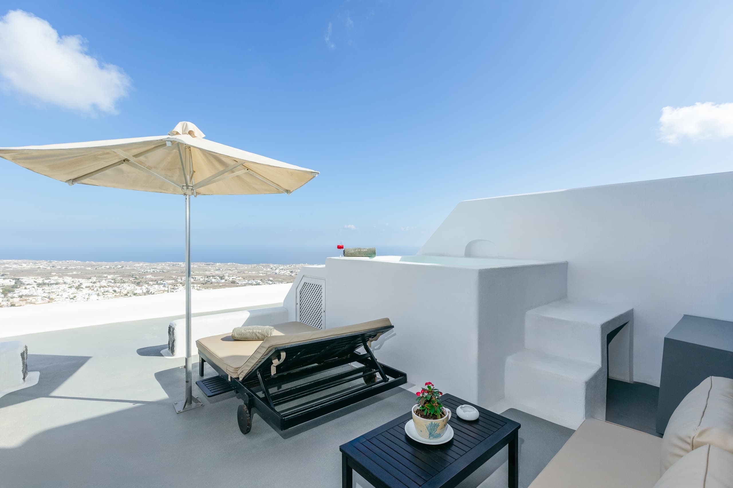 A sun lounger under a parasol on a whitewashed terrace at Carpe Diem Santorini, with the island and the sea beyond